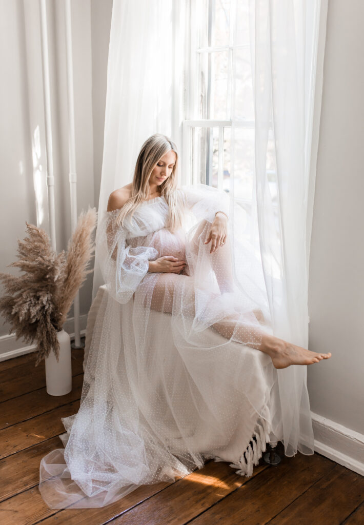 pregnant woman in sheer gown sitting in front of window by dc maternity photographer