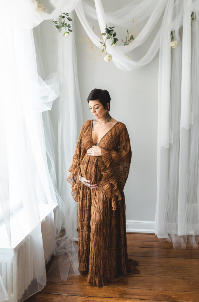 pregnant woman in brown dress holding belly in front of window by dc maternity photographer
