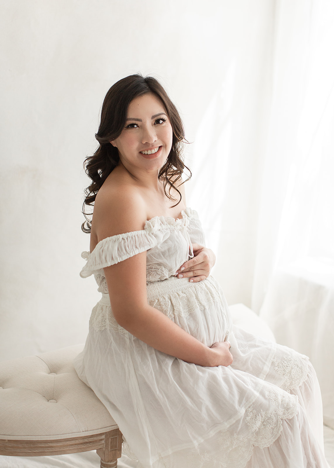 A mother to be sits by a window on an antique bench in a white maternity dress