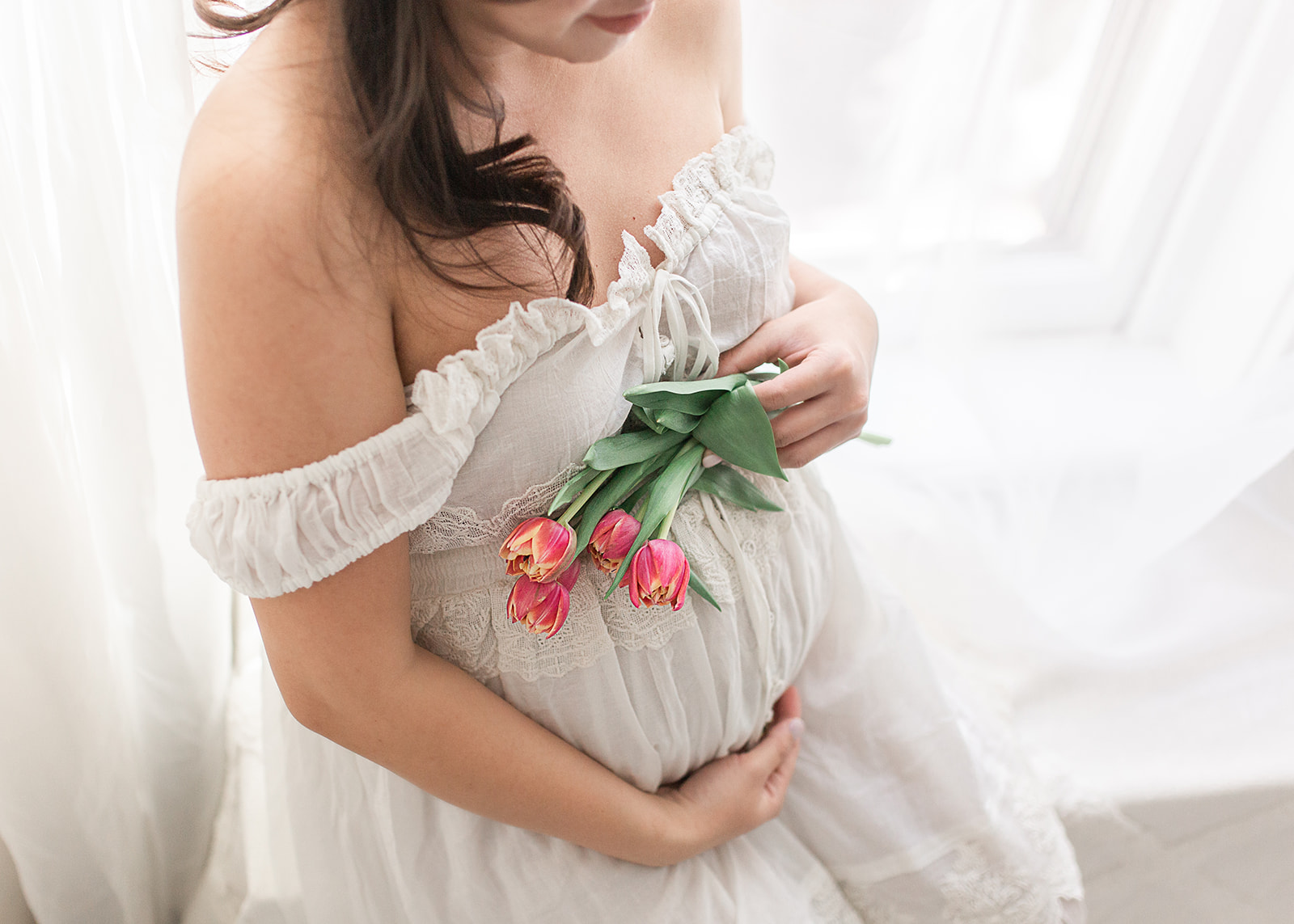 A mom to be holds a bundle of pink tulips while sitting in a window sill