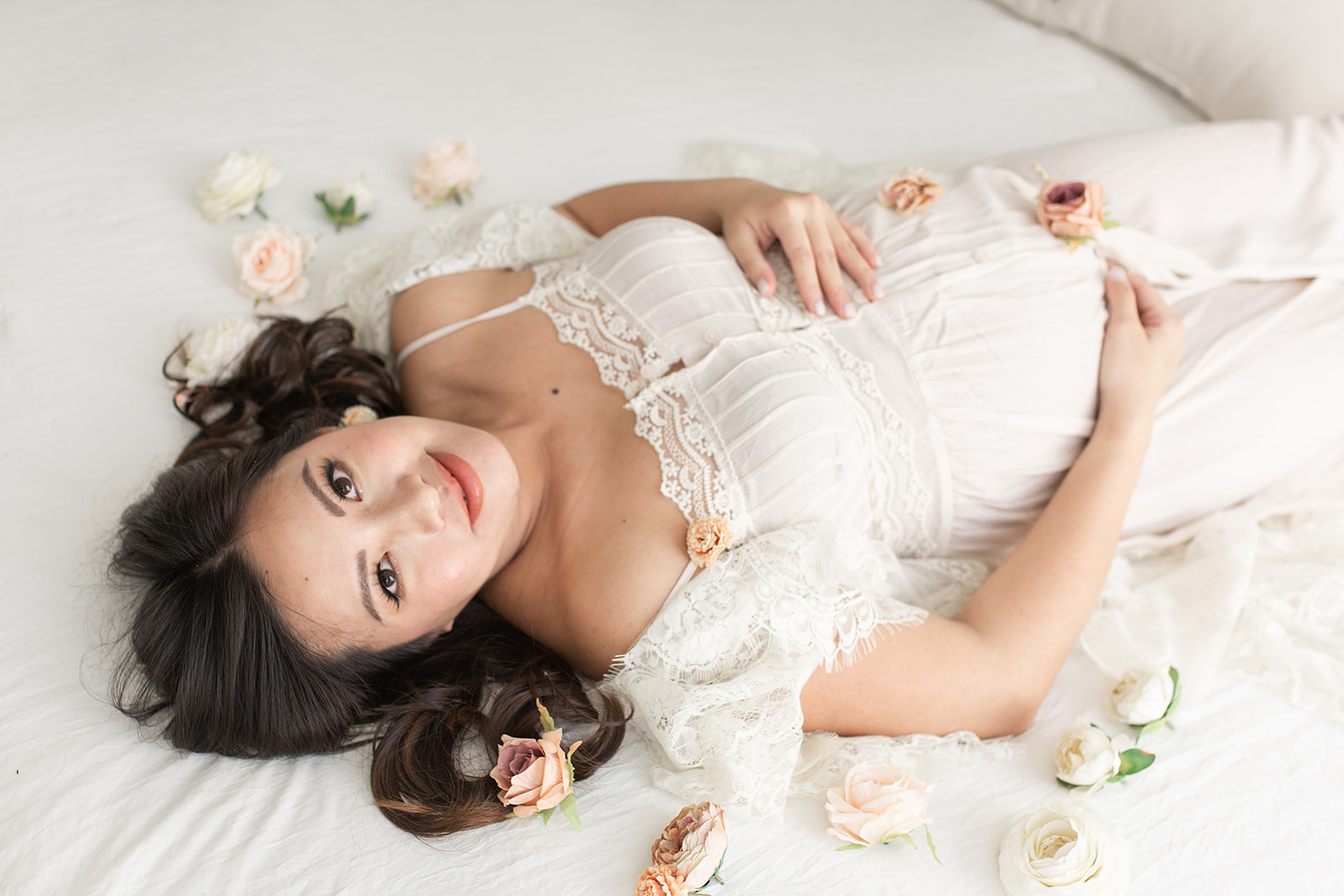 A mother to be lays on a white bed covered in carnations wearing a white maternity dress Prenatal Massage DC