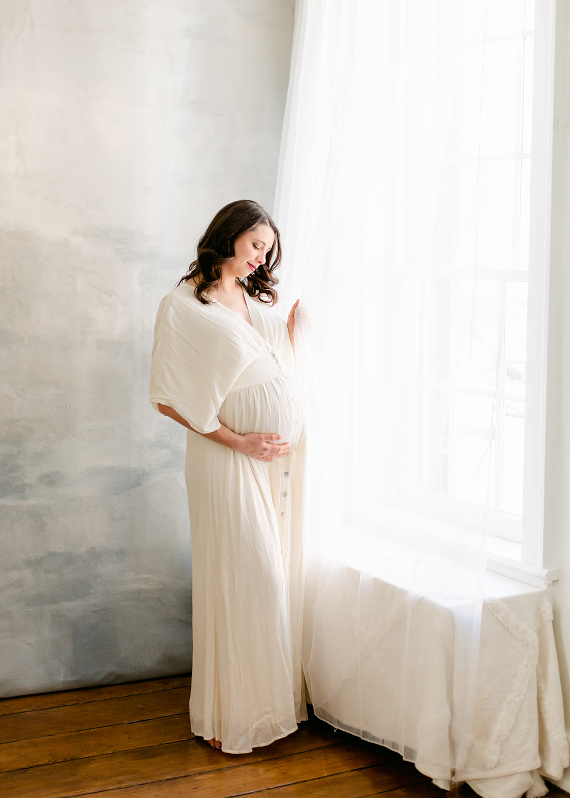 mother to be stands in a studio next to a window wearing a white maternity gown DC Doulas