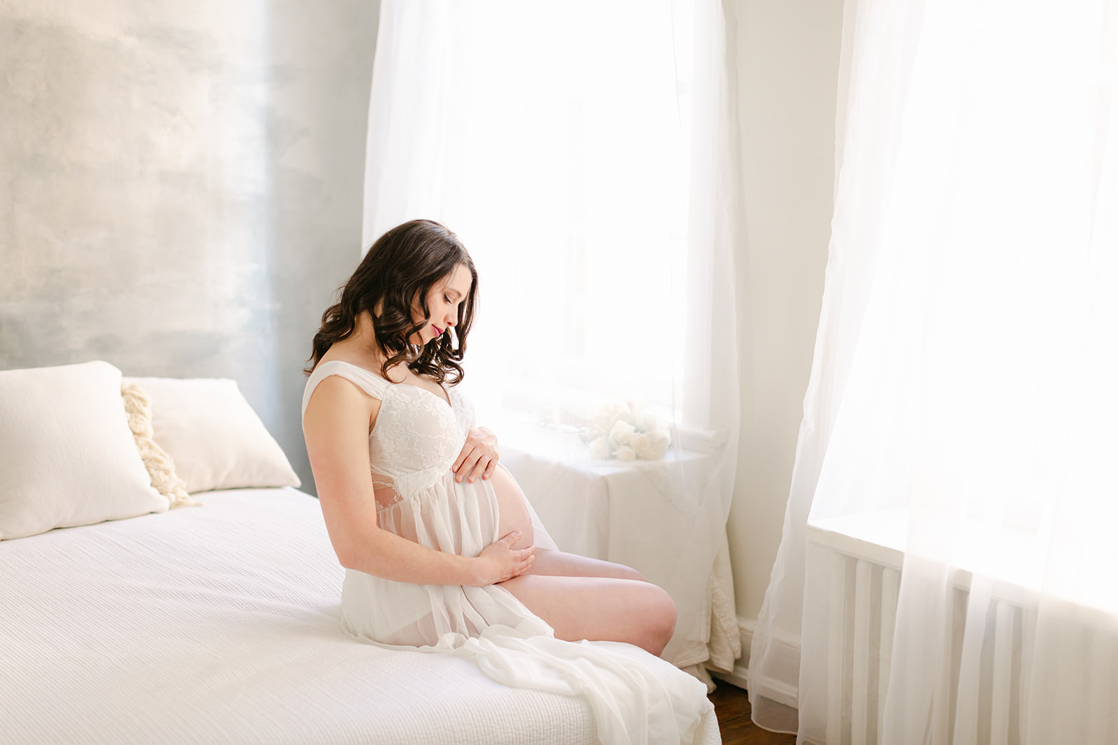 A pregnant woman sits on the edge of a bed wearing while maternity gown while looking down on her bump Prenatal Yoga DC