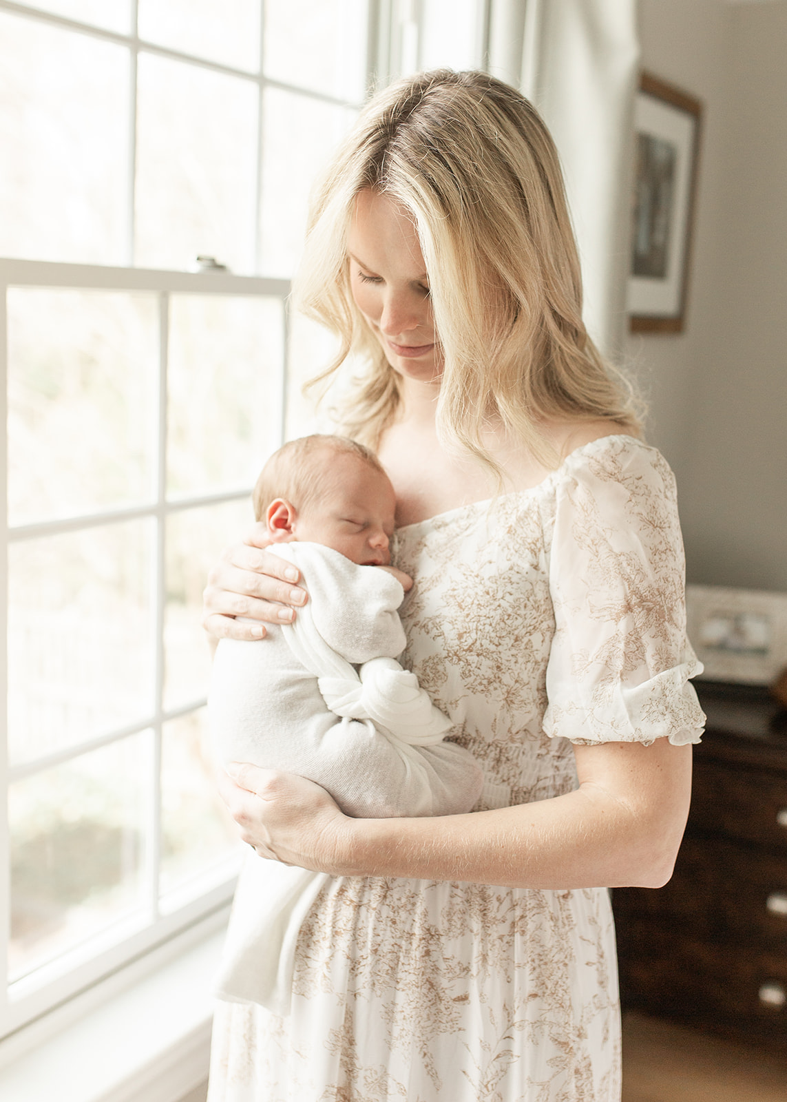 A mother stands in a white patterned dress in front of a window in a nursery holding her newborn baby who is sleeping on her chest baby boutique Washington DC