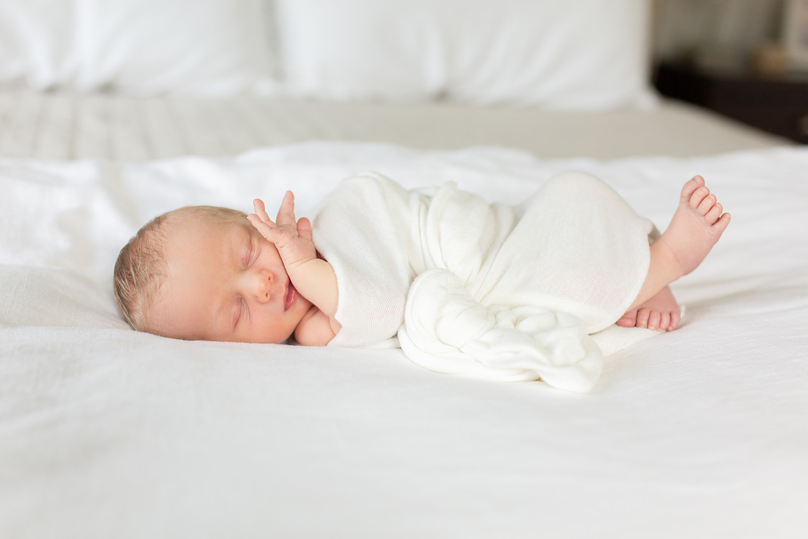 A newborn baby sleeps on a white bed wrapped in a white blanket with a hand on it's cheek