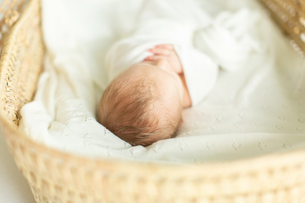A newborn baby sleep in a white swaddle with hands poking out in a wicker basket and white blankets Northern VA baby boutiques